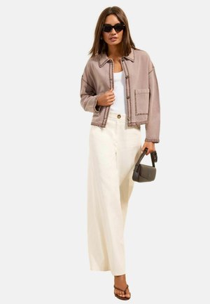 Nő bézs rövidített dzsekiben, fehér bő szárú nadrágban, fehér felsőben, fekete napszemüvegben, barna szandálban, kezében egy kis sötét táskát tart.