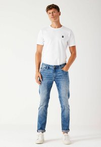 Camiseta blanca de cuello redondo con un pequeño logo negro, combinada con unos vaqueros de denim azul claro, ligeramente desgastados. Zapatillas blancas completan el atuendo.