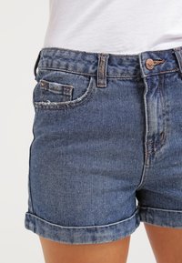 Denim-shortsit, joissa on sininen pesu, käännetyt lahkeet, viisi taskua ja metallinappisuljenta. Kankaassa on teksturoitu pinta.