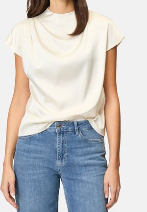 Blusa beige de manga corta hecha de tela lisa, con cuello alto y diseño drapeado, combinada con vaqueros azules clásicos.