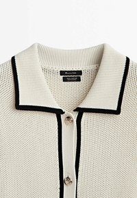 Jasnobeżowy bawełniany sweter typu polo z fakturowanym splotem, czarnymi akcentami na kołnierzyku oraz dwoma guzikami z przodu. Pod kołnierzykiem widoczne metki.