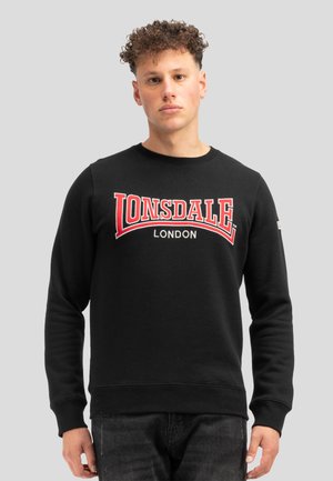 Ung mand med krøllet hår iført en sort sweatshirt med rød "LONSDALE LONDON" tekst på fronten mod en neutral grå baggrund.