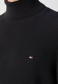 Schwarzer Rollkragenpullover aus strukturiertem Strickstoff mit einem kleinen Logoaufnäher in Rot, Weiß und Blau auf der Brust.