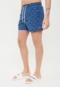 Costumi da bagno blu navy con un motivo ripetuto in verde brillante e blu. Presentano un elastico in vita con cordoncino e tasche laterali.