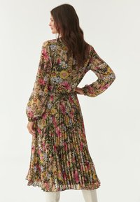 Gefaltetes Blumenkleid mit langen, transparenten Ärmeln, lebhaften pinken, gelben und blauen Blumen auf einem dunklen Hintergrund und einer definierten Taille.