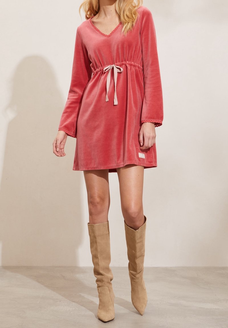 Femme portant une robe longue en velours rose poudré à manches longues, avec une taille à cordon, et des bottes en daim beige montant jusqu'aux genoux, debout sur un sol clair.