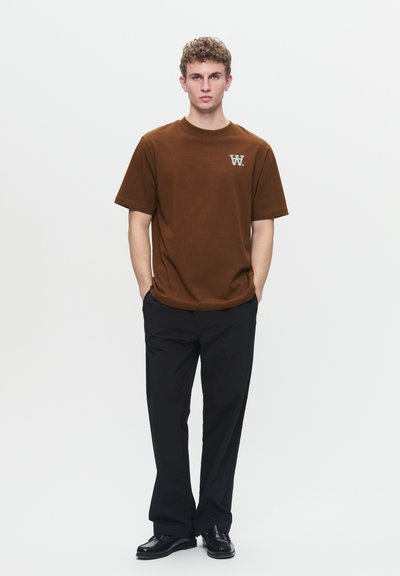 T-shirt in cotone marrone con maniche corte, con un logo di colore chiaro sul lato sinistro del petto. Indossato con pantaloni neri e scarpe nere.