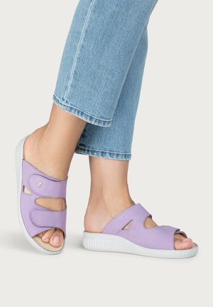 Person trägt hellblaue Jeans und fliederfarbene Slip-On-Sandalen mit offenem Zeh und weißen strukturierten Sohlen, steht vor einem einfarbigen Hintergrund.