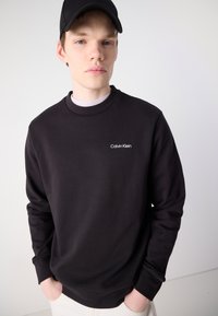 Fekete, kerek nyakú pulóver, puha anyagból készült, bal mellrészen kis, fehér "Calvin Klein" logóval. Egyszerű, testhezálló kialakítás.