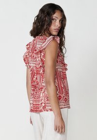 Top rosso con motivo a fantasia e scollatura arricciata, caratterizzato da design intricati. Il tessuto è leggero, abbinato a pantaloni larghi di colore chiaro.