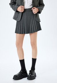 Grijze geplooide rok van textuurstof, met een aansluitende tailleband. Gecombineerd met zwarte enkelkousen en glanzende zwarte plateau schoenen.