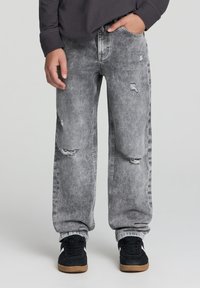 Jeans in denim grigio con vestibilità rilassata, caratterizzati da dettagli strappati e orli sfrangiati. Abbinati a sneakers nere con suola color cuoio.