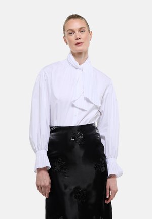 Femme portant un chemisier blanc plissé avec un col noué en forme de nœud papillon et une jupe en satin noir ornée de détails floraux, debout devant un fond uni.