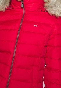 Czerwona kurtka puchowa z zapięciem na zamek, z kapturem wykończonym futerkiem, pikowaną fakturą i boczną kieszenią. Małe logo widoczne na klatce piersiowej.