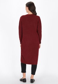 Femeie văzută din spate purtând un cardigan lung de tricot burgundiu peste pantaloni scurți negri și loafers negri, stând pe un fundal alb.