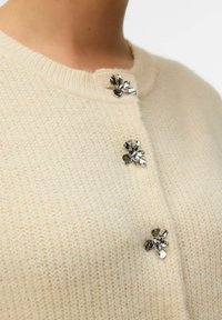 Cardigan lavorato a maglia crema con scollo rotondo e bottoni floreali argentati come chiusura, che mette in evidenza una texture morbida e una cucitura uniforme.