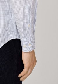 Mano masculina junto al torso, vistiendo una camisa blanca con un patrón de cuadros azules y puño abotonado, combinada con pantalones de pana negros.