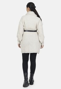 Krämbelagd ribbad stickad cardigan med hög krage, åtdragen i midjan med ett svart bälte. Bärs över svarta leggings och ankelboots.