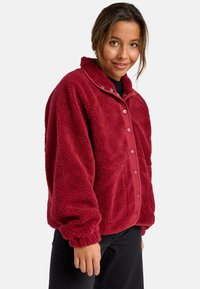 Donna che indossa una giacca di pile rossa con bottoni a pressione, pantaloni neri, sorridente, con i capelli raccolti all'indietro, su uno sfondo bianco.