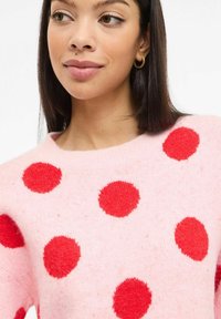 Roosa kampsun, millel on suured punased täpid. Valmistatud pehmest tekstuuriga kangast, ümara kaelusega. Lõdvalt istuv ja langevate õlgadega.