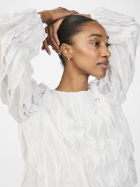 Blusa bianca con texture e maniche a volant, dal taglio morbido e una leggera lucentezza. Indossata con un semplice orecchino e capelli raccolti.