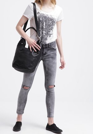 Bolso de cuero negro con textura suave. La modelo lleva una camiseta blanca con estampado gráfico, jeans grises rotos y zapatos negros con cordones.