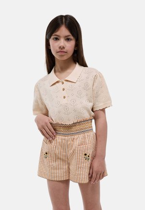 Fille portant un polo en maille crème à manches courtes et un short taille haute à carreaux orange avec broderie de tournesols sur les poches.