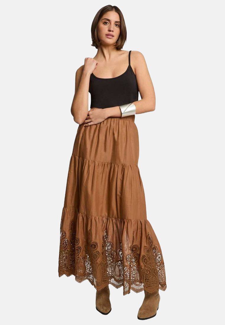 Jupe maxi brune en plusieurs niveaux, fabriquée en tissu léger avec des accents en dentelle au bas. Associée à un débardeur noir et des bottines.