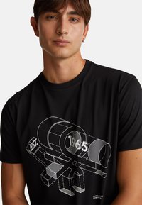 Černé bavlněné tričko s grafickým designem v bílé barvě, zahrnující geometrické tvary a text "1965". Krátké rukávy a kulatý výstřih.