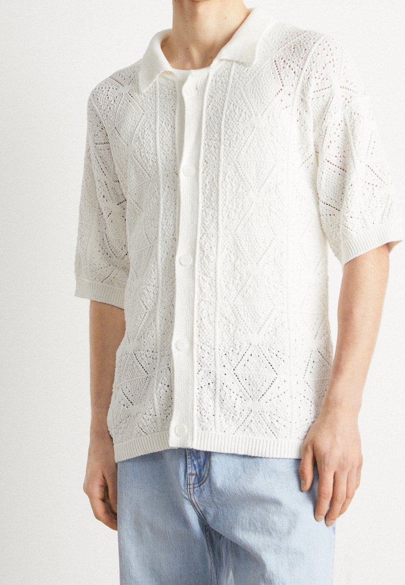 Witte gebreide cardigan met knoopsluiting en korte mouwen, met ingewikkelde diamantpatronen en een ribgebreide zoom en kraag. Draagt over lichtblauwe spijkerbroek.