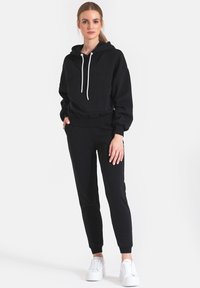 Czarna bluza z kapturem z białymi sznurkami, przednią kieszenią i dopasowanymi mankietami, zestawiona z czarnymi dresami w tym samym kolorze. Model nosi białe sneakersy.