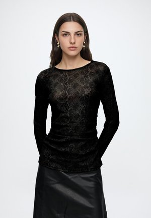 Femme aux longs cheveux bruns portant des boucles d'oreilles créoles dorées, un haut noir à manches longues à motifs et une jupe en cuir noire, debout sur fond blanc.