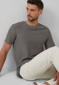 Graues kurzärmeliges T-Shirt mit geripptem Textur, Rundhalsausschnitt, kombiniert mit hellen Jeans, vor einem neutralen Hintergrund.