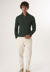 Dunkelgrüner Strickpullover mit einem Zopfmuster und einem Reißverschlusskragen, kombiniert mit cremefarbenen Hosen und hellbraunen Schuhen.