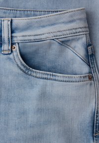 Gros plan sur la poche d'un jean en denim bleu clair avec coutures visibles, passants de ceinture et rivets métalliques sur un tissu texturé.