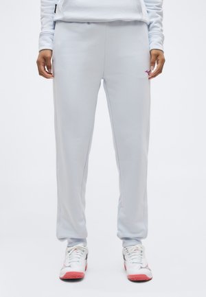 Άτομο που φορά ανοιχτό γκρι παντελόνι φόρμας και λευκά αθλητικά παπούτσια με ροζ σόλες, στέκεται μπροστά σε μια απλή λευκή κουρτίνα.