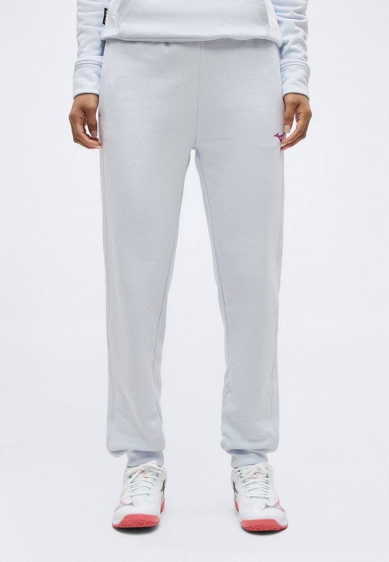 Άτομο που φορά ανοιχτό γκρι παντελόνι φόρμας και λευκά αθλητικά παπούτσια με ροζ σόλες, στέκεται μπροστά σε μια απλή λευκή κουρτίνα.