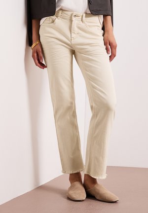 Frau trägt cremefarbene, gerade geschnittene Jeans mit ausgefransten Säumen, braune Slip-On-Schuhe, schwarze Jacke und goldenes Armband, steht vor einer weißen Wand.