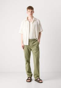 Lightweight, striped short-sleeve shirt in cream, paired with loose olive green pants. The model wears dark brown sandals, standing on a white background.
