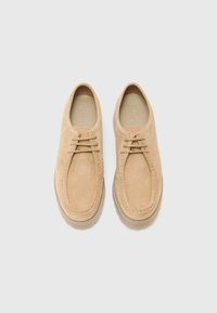 Chaussures en daim beige avec un bout arrondi, des coutures décoratives et des lacets. La semelle est texturée et en caoutchouc, offrant adhérence et durabilité.