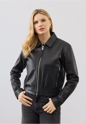 Frau mit langen blonden Haaren trägt eine schwarze Lederjacke mit Reißverschluss und dunkle Jeans, steht vor einem schlichten hellgrauen Hintergrund.