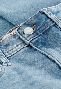 Los jeans de mezclilla azul claro presentan un botón plateado con texto grabado, costuras visibles y una textura ligeramente desgastada.
