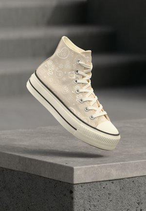Basket montante beige avec motifs cachemire blancs et semelle épaisse, flottant au-dessus d'un rebord en béton gris sur un fond sombre flou.