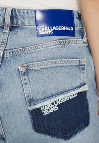 Modré džínové kalhoty s modrou koženou nášivkou a kontrastní tmavě modrou zadní kapsou s bílým vyšívaným textem. Jsou viditelné třepené okraje.