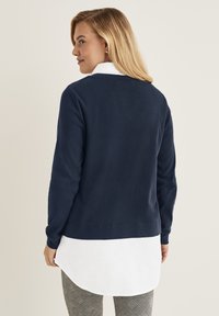 Marinblå tröja med ribbad textur, lagd över en vit skjorta med krage, stylad med mönstrade grå byxor.