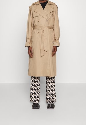 Osoba ubrana w beżowy płaszcz trencz do kolan z paskiem, czarną bluzkę, wzorzyste czarno-białe spodnie o szerokich nogawkach oraz czarne buty z kwadratowym noskiem.