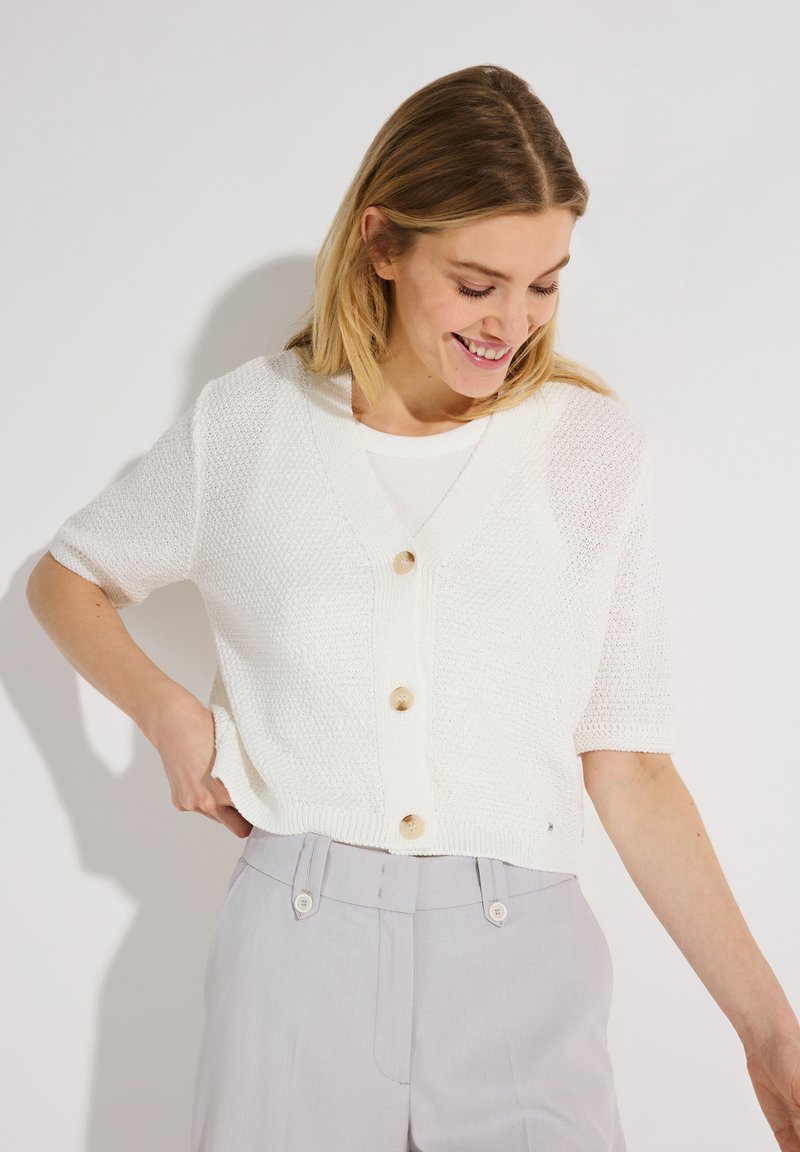 Woman smiling while looking down, wearing a white short-sleeve knit cardigan with buttons and light gray high-waisted pants.