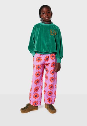 Grüner Samt-Sweatshirt mit floralem Stickmuster, rosa gemusterte gesteppte Hose mit roten und schwarzen Blumen sowie braunen Schnürschuhen.
