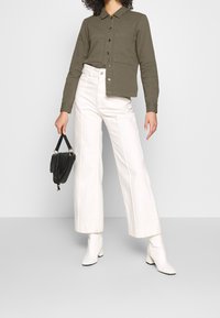 Olivově zelená zkrácená košile s knoflíky a dvěma kapsami na hrudi, kombinovaná s vysokými bílými širokými kalhotami a bílými kotníkovými botami. Černá kabelka v ruce.