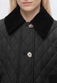 Femme portant une veste matelassée noire avec un large col en velours côtelé et des boutons argentés gravés avec "Barbour."
